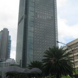 Ayala Tower One is a skyscraper with facade made of square glass panels reflecting a cloudy sky, surrounded by palm trees and a busy street. This is the tower as seen from sidewalk in front of Allied Bank Center.