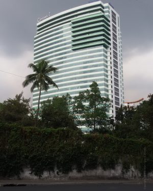 The Cyberpark Tower 2 is a rectangular building with a curving front and back surface. Its facade is made of stripes of alternating rows of white concrete and green glass panel windows. On the upper part on the right side is a protruding square section that seems like several levels of verandas. This picture was taken from EDSA cor. Tuazon Boulevard. A white wall with plants crawling on it obscures the lower levels of Cyberpark Tower 2.