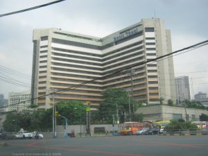 The facade of Dusit Thani Manila features a pattern of horizontal stripes of concrete walls and indented glass panels. It is surrounded by a concrete barrier that has greenery growing within it. This is the hotel as seen from opposite corner on EDSA northbound side.