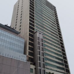 IBM Plaza has a tall blocky appearance and has a facade of mostly gray concrete. It also has what looks like an external elevator shaft that reaches halfway to the floors above the podium. This picture shows the back of IBM Plaza, as seen from the compound of St. Pio of Pietrelcina Chapel.