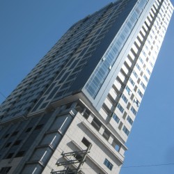 The lower podium floors of Mayfair Tower have a beige concrete exterior. In contrast, the upper floors feature white concrete and blue glass windows. The photograph was taken from the sidewalk along UN Avenue near the opposite corner.
