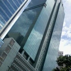 Petron Megaplaza, a modern glass skyscraper reflecting the surrounding cityscape under a cloudy sky shot from front of ECC (Employment Compensation Commission) Building. The partially visible building on the upper-left is BPI Buendia Center.