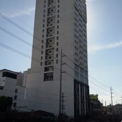 The Privato Ortigas Hotel is a high-rise building with small square glass windows on its upper 2/3. Its lower 1/3 with a larger area has a branching plant-like design in front, which seems to be made of metal. The side of the building visible in this photo has recesses at every floor on the upper 2/3 containing what appears to be external parts of airconditioning units. The side of the lower 1/3 is a flat concrete wall as if it's a firewall.
