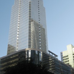 A tall skyscraper with a glass facade, set against a clear blue sky, with a wider podium structure and trees at its base.