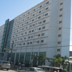 Santa Ana Hospital is a low-rise rectangular building with a facade of white concrete, green glass panels, and several small windows. This picture was taken from the sidewalk on the opposite side.