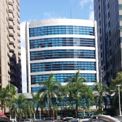 Suntree Tower is a cuboid structure atop a shorter cuboid. Its facade is that of gray tiles and blue glass panels. This is Suntree, seen from along Garnet Road with a parking space between it and Sapphire Road.