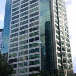 The Taipan Place is a tall and narrow cuboid building with a facade of blue glass panels, gray tiles, and dark marble. This is The Taipan Place, as seen from the sidewalk on the opposite side of Emerald Avenue.