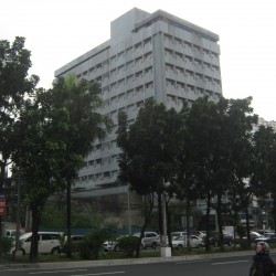 The building features a straightforward cuboid structure with a gray concrete exterior and evenly spaced windows. The shot was taken from the sidewalk on the southbound side of Quezon Ave.