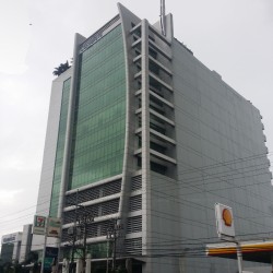 Two Sanparq as seen from across the Shell gasoline station adjacent to it. It has a large sign that says “Two Sanparq” on the top floor. The image also shows some cars and motorcycles parked in front of the tower, as well as some trees and plants on the sidewalk. The image was taken from a low angle, giving a sense of height and perspective to the tower.